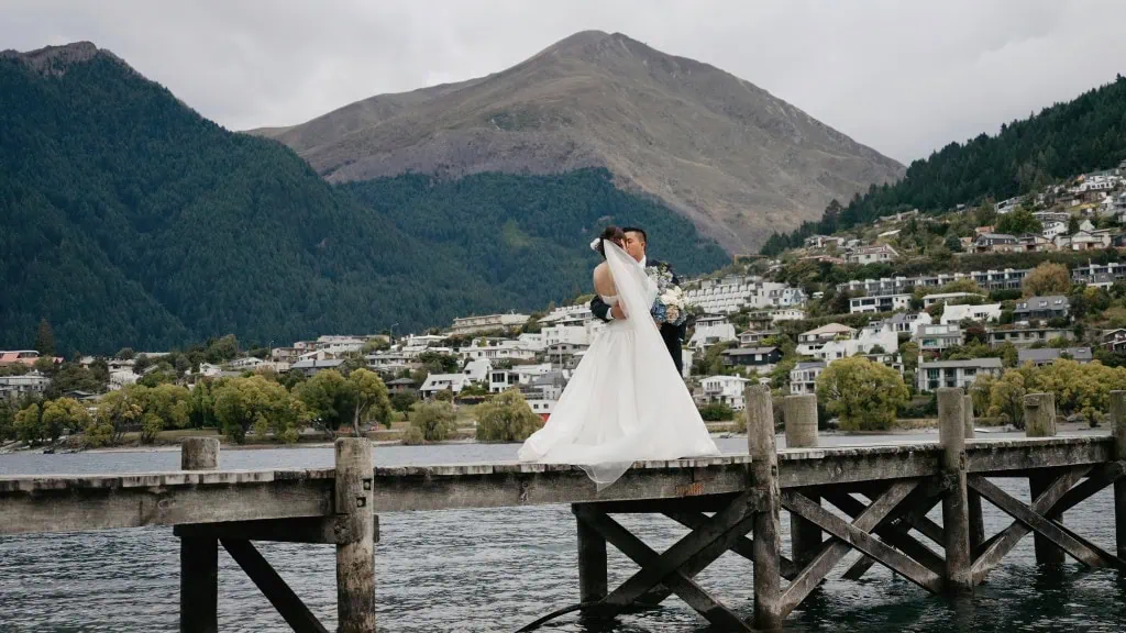 queenstown new zealand heli wedding elopement photographer videographer | Hitomi and Winston stand on a wooden dock over a serene body of water, embracing. Behind them, the majestic Earnslaw Burn streamlines through green hills and mountains, with a quaint town nestled into the landscape—a perfect setting for their heli-wedding.