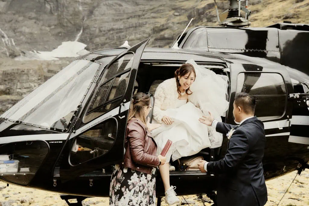 queenstown new zealand heli wedding elopement photographer videographer | At an Earnslaw Burn Heli-Wedding, bride Hitomi steps out of a black helicopter with the assistance of two people. Smiling and wearing a white dress, she holds Winston’s hand while the other person supports her from behind.