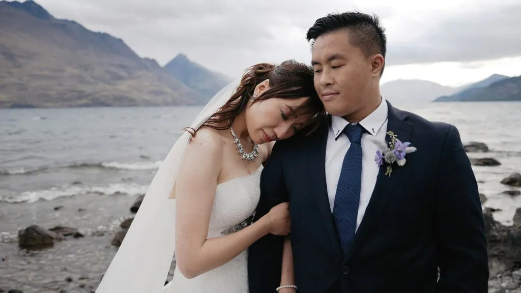queenstown new zealand heli wedding elopement photographer videographer | A bride in a white dress and veil rests her head on the groom's shoulder by a lakeshore, with mountains and cloudy skies in the background. The groom wears a dark suit and a flower boutonniere. This picturesque scene captures the romance of an Earnslaw Burn Heli-Wedding for Hitomi & Winston.