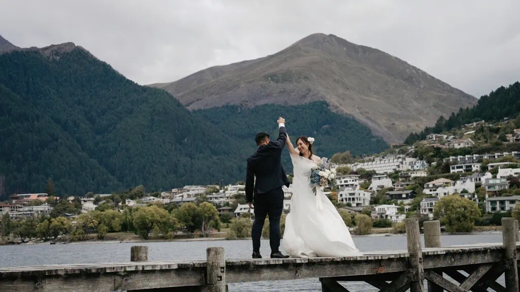 queenstown new zealand heli wedding elopement photographer videographer | Hitomi & Winston dance on a wooden dock by a serene lake, with majestic mountains and a quaint town in the background—a perfect setting for their unforgettable Earnslaw Burn Heli-Wedding.