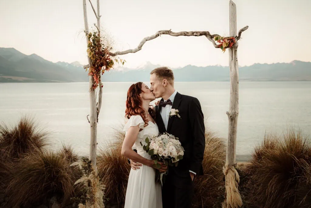 queenstown new zealand heli wedding elopement photographer videographer | A couple in wedding attire kisses under a rustic wedding arch, with a scenic lake and mountain landscape in the background. Captured by a Queenstown wedding photographer, the bride holds a bouquet.