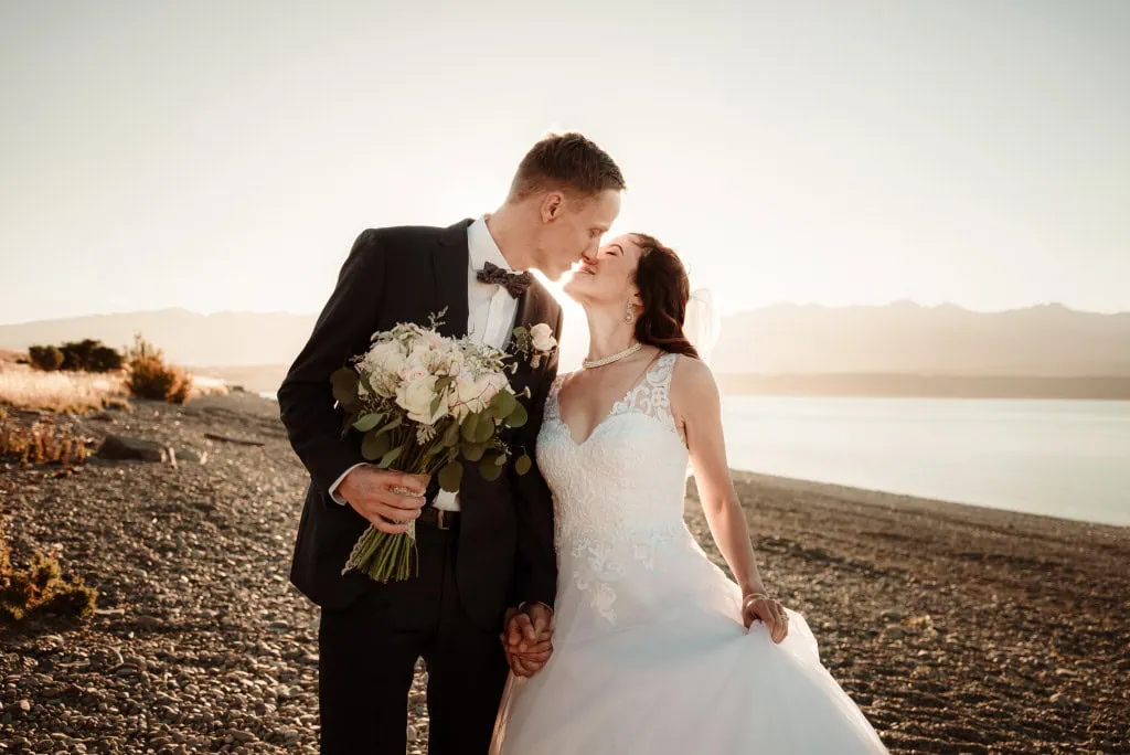 queenstown new zealand heli wedding elopement photographer videographer | A bride and groom kiss while holding hands on a rocky beach at sunset. The groom, clutching a bouquet of flowers, seems to be savoring the moment captured by their talented Queenstown wedding photographer.