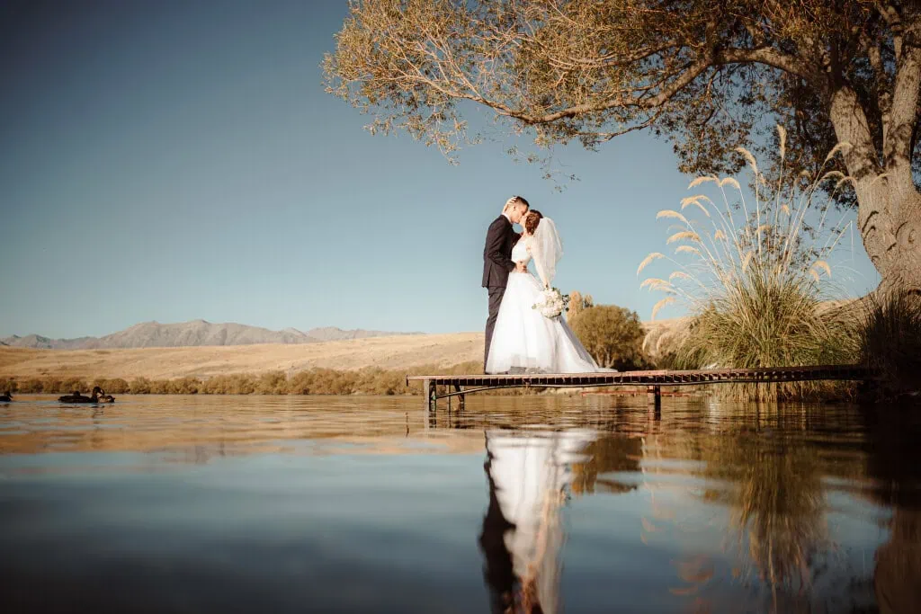 queenstown new zealand heli wedding elopement photographer videographer | A bride and groom stand on a wooden dock over a calm body of water, embracing under a tree. Captured by a talented Queenstown wedding photographer, the backdrop features a dry, open landscape with hills under a clear sky.