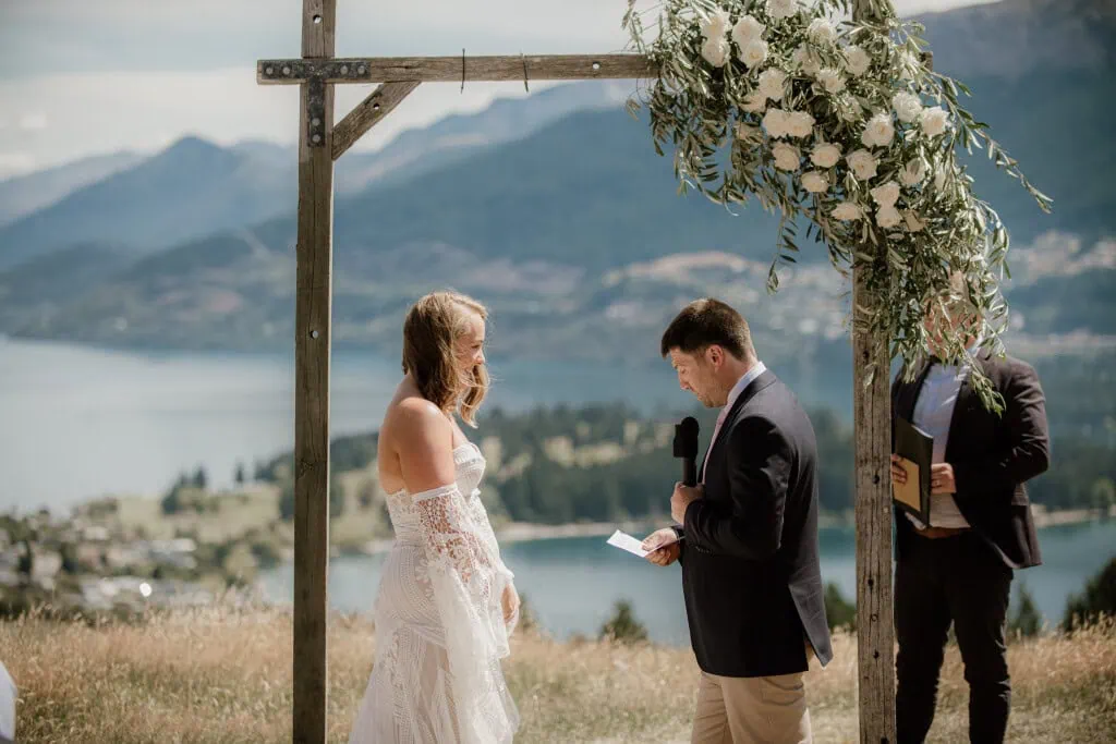 queenstown new zealand heli wedding elopement photographer videographer | A couple stands under a wooden arch adorned with white flowers during an outdoor wedding ceremony at Deer Park Heights in Queenstown, overlooking a lake and mountains, with the groom reading from a piece of paper.