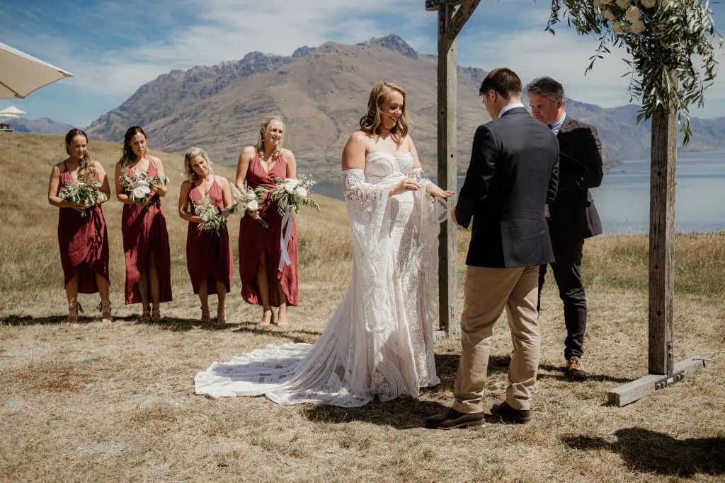 queenstown new zealand heli wedding elopement photographer videographer | A couple exchanges rings during their wedding ceremony at stunning Deer Park Heights in Queenstown, with bridesmaids in red dresses and a breathtaking mountainous landscape in the background.