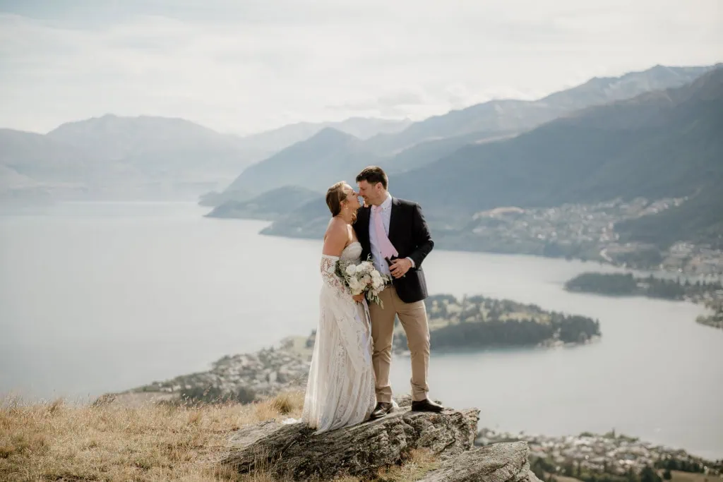 queenstown new zealand heli wedding elopement photographer videographer | A couple in wedding attire share a kiss on a rocky outcrop at Deer Park Heights, with the stunning backdrop of Queenstown's lake and mountainous terrain.