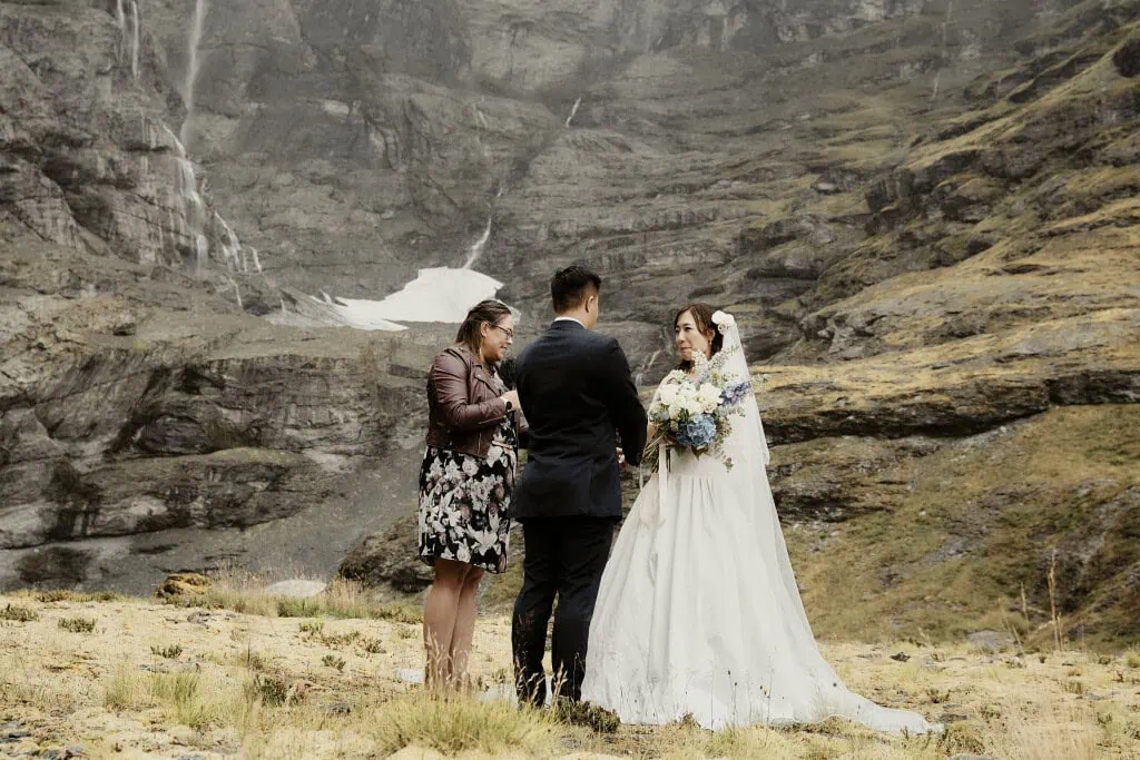 queenstown new zealand heli wedding elopement photographer videographer | A couple stands in wedding attire with an officiant in front of the rocky, mountainous backdrop of Earnslaw Burn during an unforgettable heli-wedding ceremony.