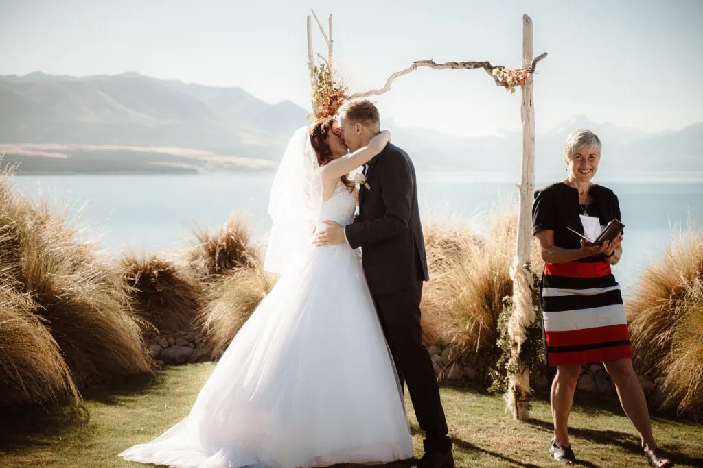 queenstown new zealand heli wedding elopement photographer videographer | A couple shares a kiss at their intimate elopement wedding by Lake Tekapo, with the majestic Mt Cook as a backdrop. Under a beautifully decorated arch, an officiant stands to the side, smiling and holding a book.