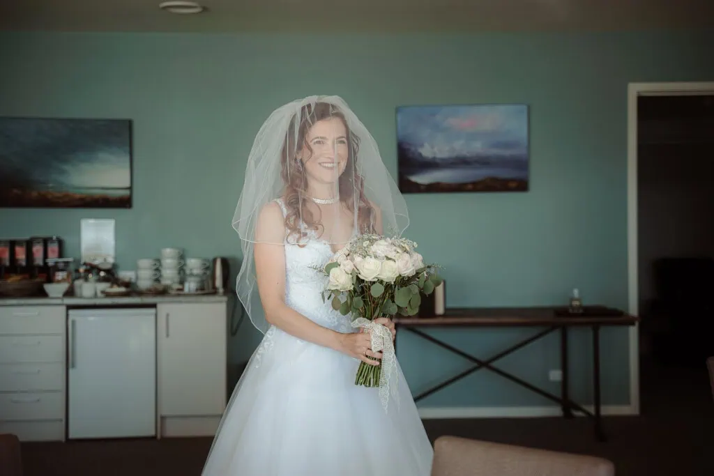 queenstown new zealand heli wedding elopement photographer videographer | A bride stands indoors wearing a white wedding dress and veil, holding a bouquet of flowers. Behind her, there is a white cupboard, paintings on the wall, and a table with various items, capturing the charm of an intimate NZ elopement wedding near Mt Cook.