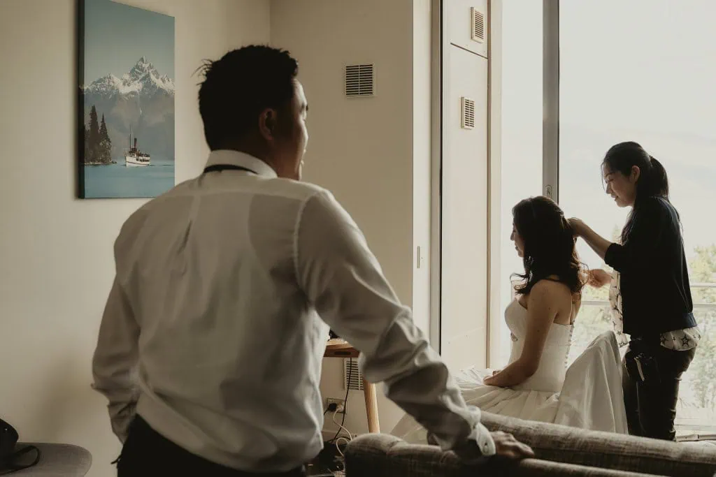 queenstown new zealand heli wedding elopement photographer videographer | A woman is sitting on a chair getting her hair styled by another woman standing behind her. A man in a white shirt, perhaps Winston, stands nearby, looking towards them intently. On the wall hangs a painting of Earnslaw Burn, adding to the room's serene ambiance.