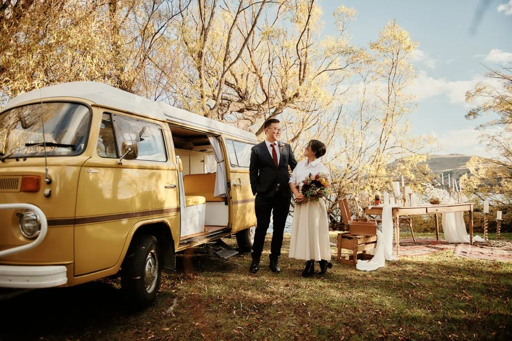 queenstown new zealand heli wedding elopement photographer videographer | A couple stands beside a yellow vintage van in a wooded area during fall, capturing the charm of their autumn elopement in Arrowtown. They are dressed formally; the man in a suit and the woman in a bridal gown holding a bouquet. A decorated outdoor table is in the background.