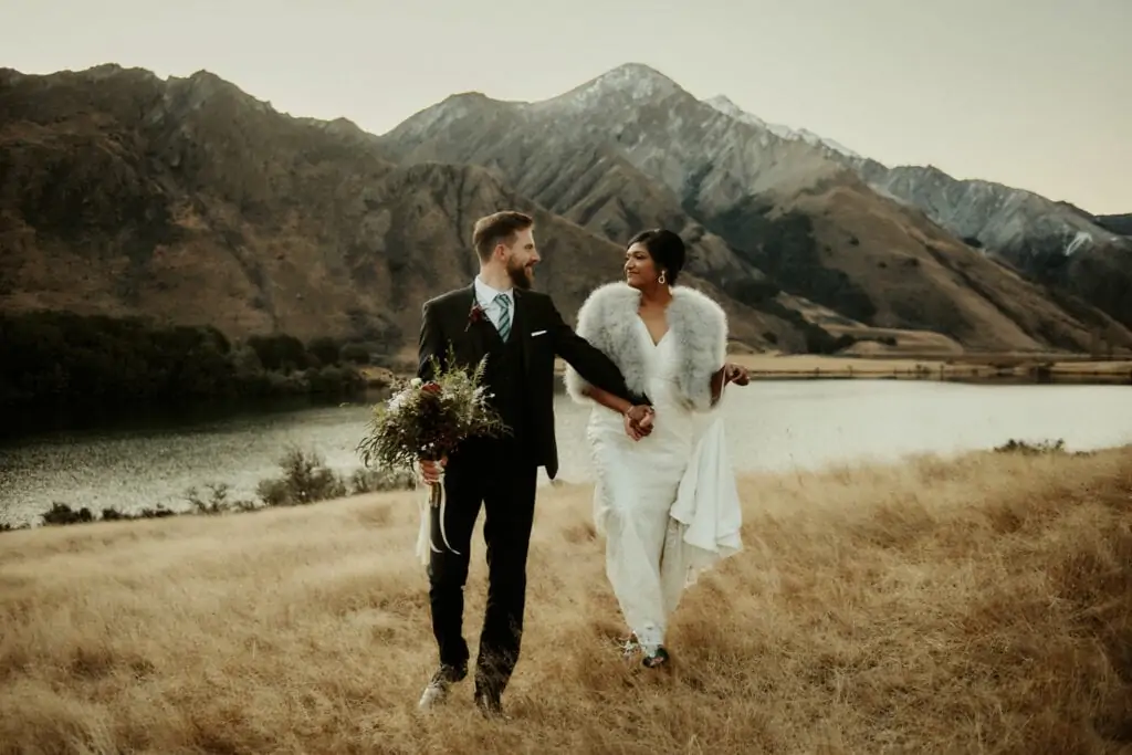 A couple enjoying their queenstown heli-wedding package by walking through a grassy field with mountains in the background.