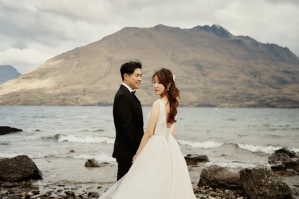 queenstown new zealand heli wedding elopement photographer videographer | A bride and groom stand on a rocky shoreline in Queenstown, NZ, with mountains in the background. The bride wears a white gown, and the groom wears a black suit. They are looking back towards the camera during their Heli Pre-Wedding Shoot.