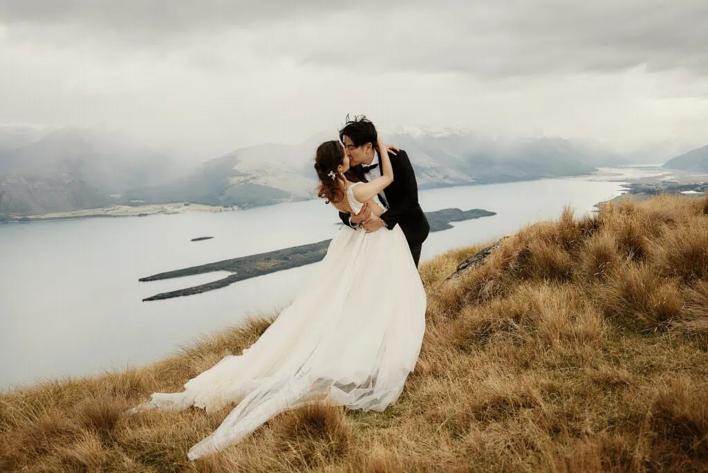 queenstown new zealand heli wedding elopement photographer videographer | Newlyweds Celine & Philip embrace and kiss on a grassy hilltop in Queenstown, NZ, with a scenic view of the lake and distant mountains under a cloudy sky during their heli pre-wedding shoot.