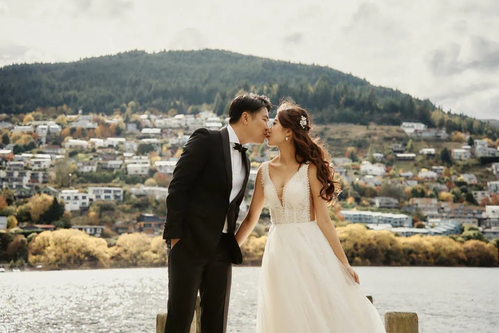 queenstown new zealand heli wedding elopement photographer videographer | A couple in formal attire kisses in front of a scenic backdrop with a lake and hillside town, capturing the magic of their Heli Pre-Wedding Shoot in picturesque Queenstown, NZ.