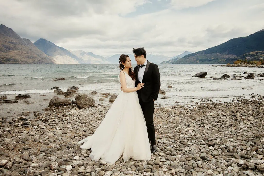 queenstown new zealand heli wedding elopement photographer videographer | Celine & Philip, dressed in wedding attire, stand on a rocky beach with a scenic view of Queenstown's mountains and lake in the background. She is wearing a white gown, and he is in a black suit with a bow tie, capturing their Heli Pre-Wedding Shoot moment.
