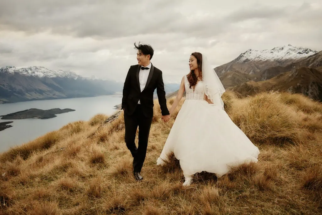 queenstown new zealand heli wedding elopement photographer videographer | A couple in wedding attire stands holding hands on a grassy hillside with a view of New Zealand's Queenstown mountains and lake in the background, capturing the essence of their Heli Pre-Wedding Shoot.