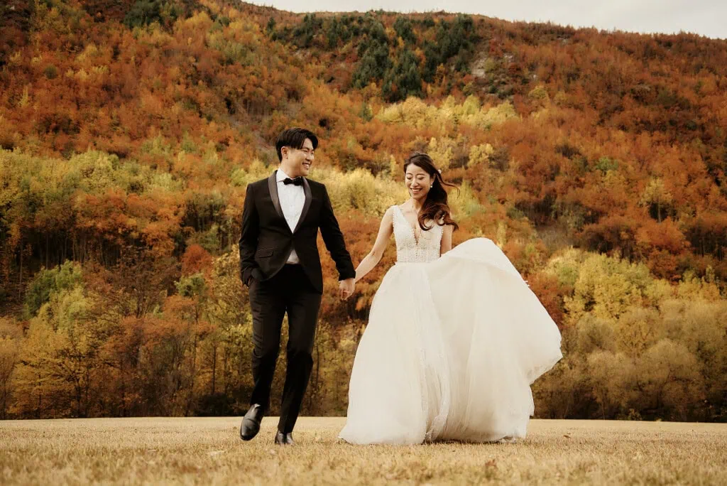 queenstown new zealand heli wedding elopement photographer videographer | A couple in wedding attire holds hands and smiles while walking on a grassy field with a colorful autumn forest in the background, capturing the essence of their Heli Pre-Wedding Shoot in Queenstown, New Zealand.