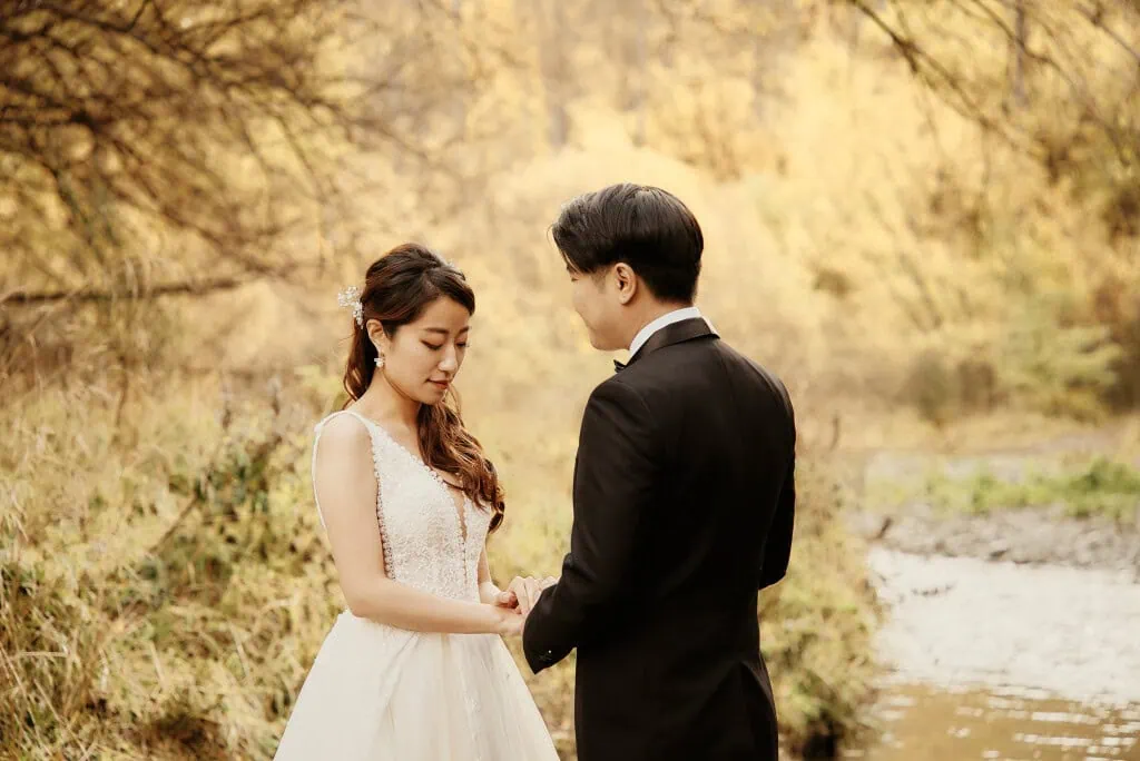 queenstown new zealand heli wedding elopement photographer videographer | A couple dressed in wedding attire stands facing each other holding hands in an outdoor setting with the autumn foliage of Queenstown, NZ, as a breathtaking backdrop for their pre-wedding shoot.