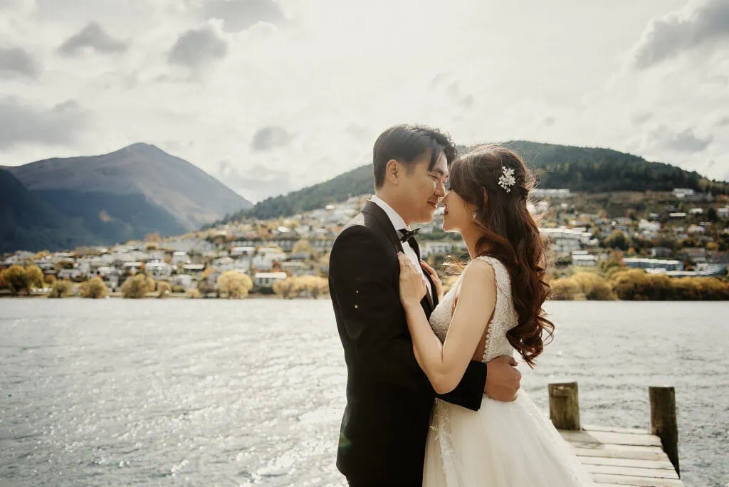 queenstown new zealand heli wedding elopement photographer videographer | Celine & Philip, in elegant wedding attire, embrace on a wooden dock by a serene lake, with the majestic mountains and charming houses of Queenstown NZ in the background under a partly cloudy sky—captured perfectly in their Heli Pre-Wedding Shoot.
