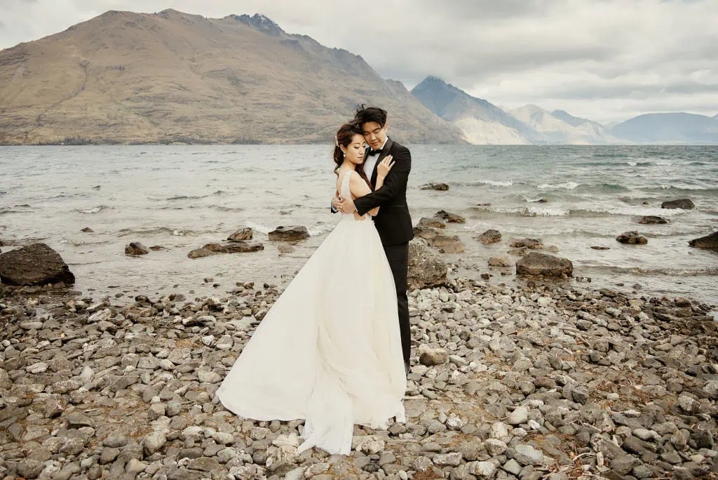queenstown new zealand heli wedding elopement photographer videographer | A couple dressed in wedding attire stands on a rocky shoreline in Queenstown, NZ, with mountains and a lake in the background. The bride wears a white gown, and the groom wears a black suit. They embrace while facing the camera during their pre-wedding shoot.