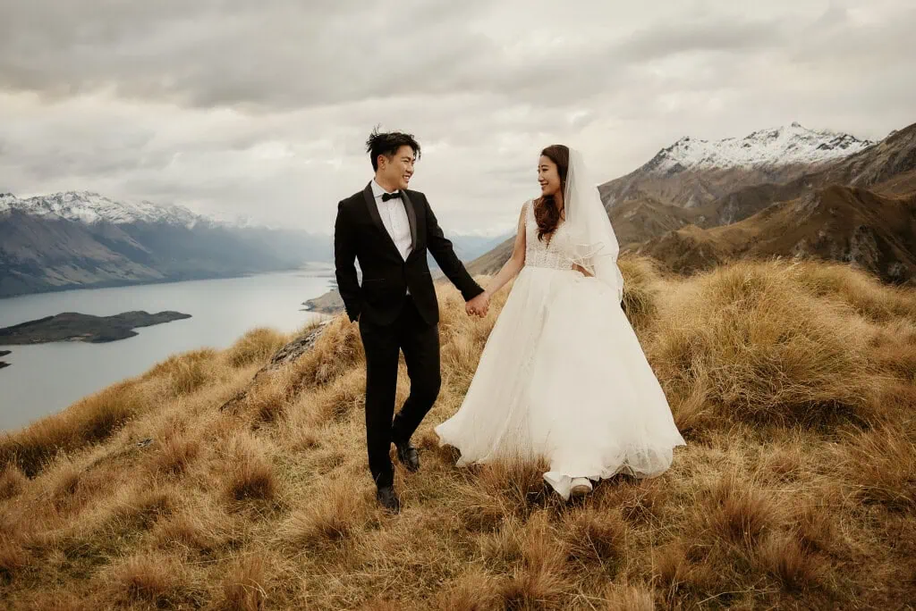 queenstown new zealand heli wedding elopement photographer videographer | A bride and groom in wedding attire walk hand in hand on a grassy hilltop, with a lake and snowy mountains in the background during their Queenstown heli pre-wedding shoot.