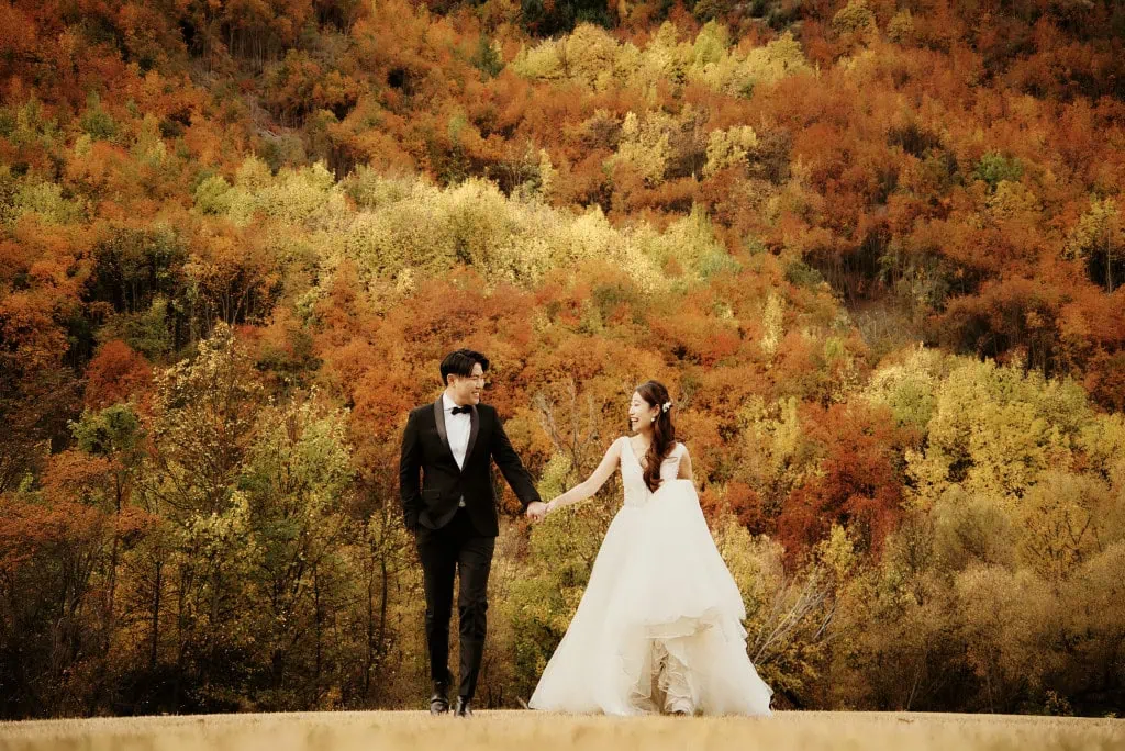 queenstown new zealand heli wedding elopement photographer videographer | A couple dressed in formal wedding attire hold hands and walk in front of a backdrop of autumn foliage in a forest during their pre-wedding shoot in Queenstown.