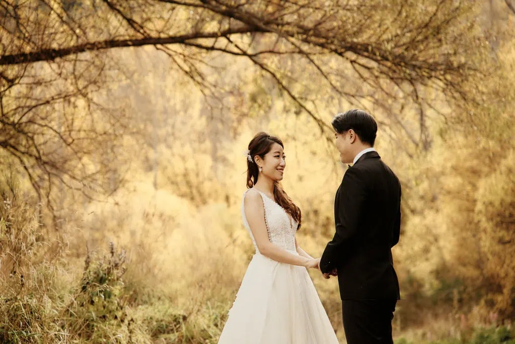 queenstown new zealand heli wedding elopement photographer videographer | A bride and groom stand facing each other and holding hands outdoors in Queenstown, surrounded by autumn trees with golden leaves—capturing the perfect moment for their pre-wedding shoot.