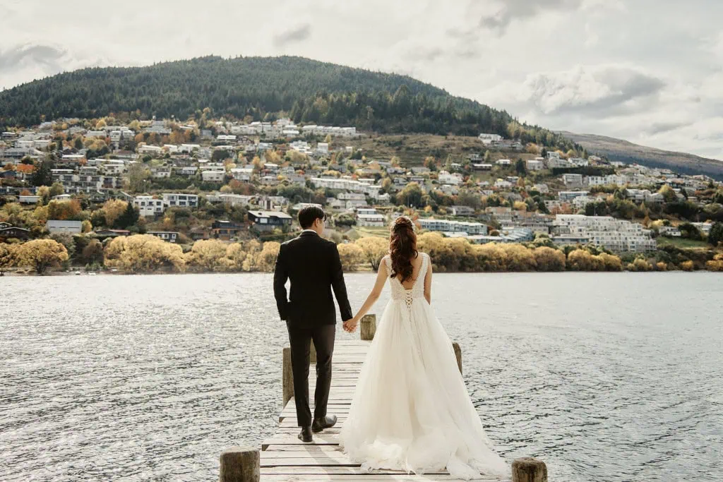 queenstown new zealand heli wedding elopement photographer videographer | A couple in wedding attire stands hand in hand at the end of a dock, overlooking a large body of water with Queenstown's hillside town in the background. Their Heli Pre-Wedding Shoot captures the breathtaking beauty of NZ's stunning landscape.