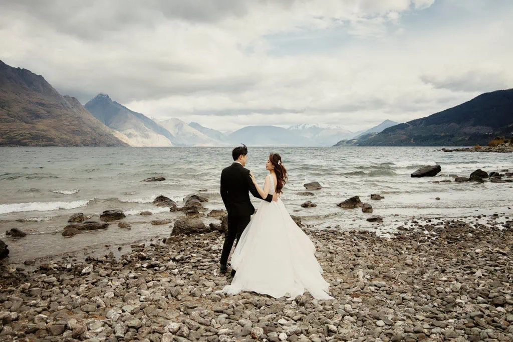 queenstown new zealand heli wedding elopement photographer videographer | A couple in wedding attire stands on a rocky shore in Queenstown, NZ, facing a mountainous landscape with a lake. The bride wears a white dress while the groom is in a black suit, capturing the essence of their Heli Pre-Wedding Shoot.