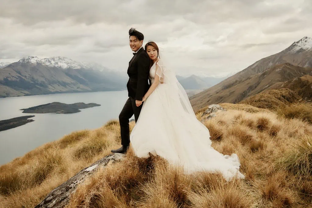 queenstown new zealand heli wedding elopement photographer videographer | A couple stands outdoors in wedding attire on a rocky hilltop near Queenstown, with expansive views of a lake and mountains in the background. The bride wears a white dress, and the groom is in a dark suit, capturing the perfect moment during their NZ Heli Pre-Wedding Shoot.