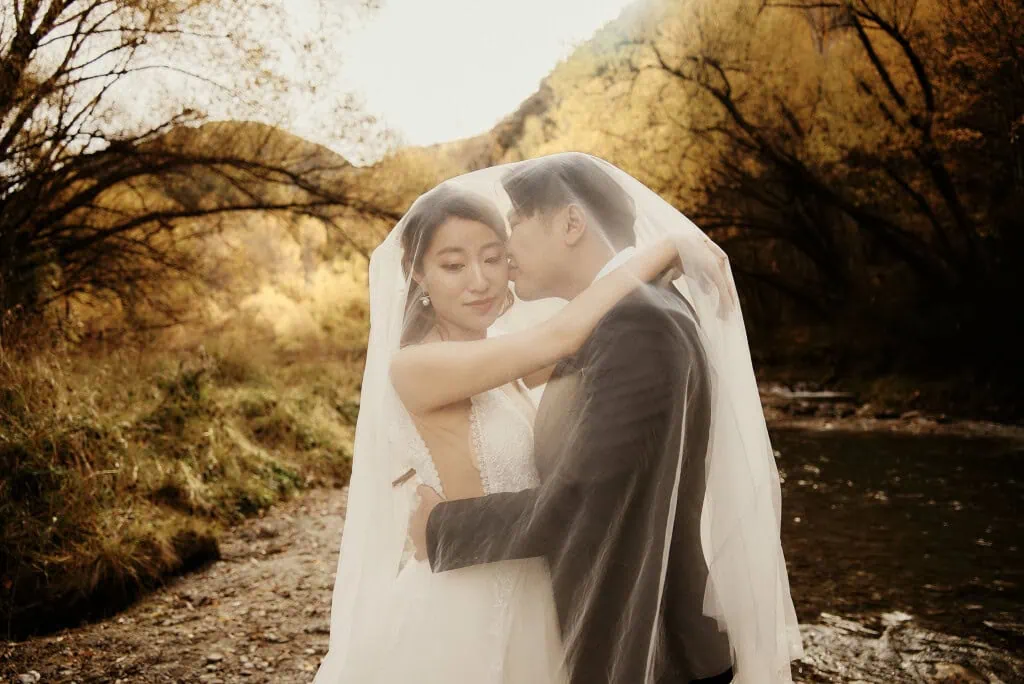 queenstown new zealand heli wedding elopement photographer videographer | A bride and groom embrace under a veil beside a narrow stream, with autumn trees in the background, during their Heli Pre-Wedding Shoot in Queenstown, NZ.