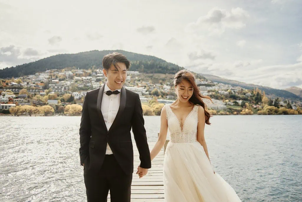 queenstown new zealand heli wedding elopement photographer videographer | A couple dressed in wedding attire holds hands while walking on a wooden pier overlooking a lake with a hilly town in the background, capturing their perfect pre-wedding shoot moment in picturesque Queenstown, NZ.