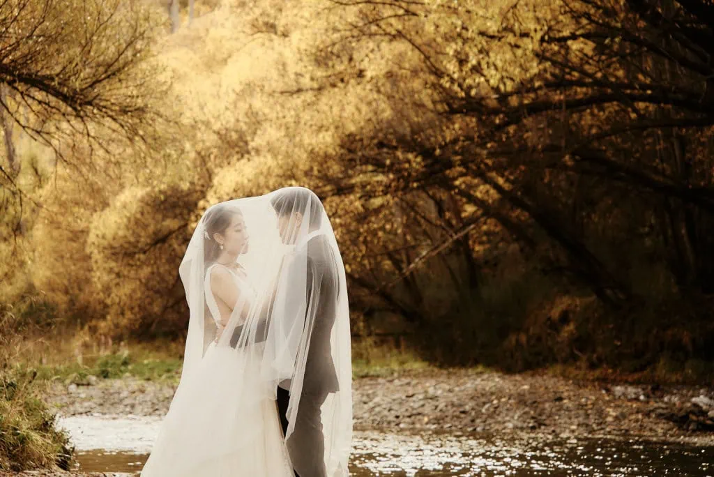 queenstown new zealand heli wedding elopement photographer videographer | Celine & Philip stand under a veil near a stream with autumn trees in the background during their Heli Pre-Wedding Shoot in Queenstown, NZ. The couple faces each other, both dressed in formal wedding attire.