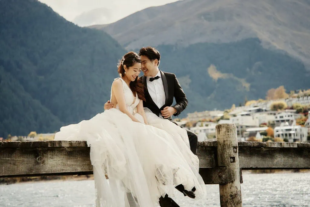queenstown new zealand heli wedding elopement photographer videographer | A bride and groom sit closely on a wooden dock in Queenstown, NZ, overlooking a serene body of water with mountains and buildings in the background. They appear joyful and relaxed in their formal wedding attire during their Heli Pre-Wedding Shoot.