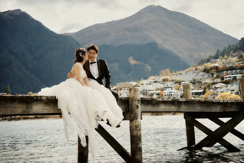 queenstown new zealand heli wedding elopement photographer videographer | A bride and groom sit on a wooden pier by a lake with the stunning Queenstown mountains and a small town in the background, capturing a perfect moment before their heli pre-wedding shoot. The bride wears a white gown, and the groom is in a black suit, embodying timeless elegance against NZ's natural beauty.