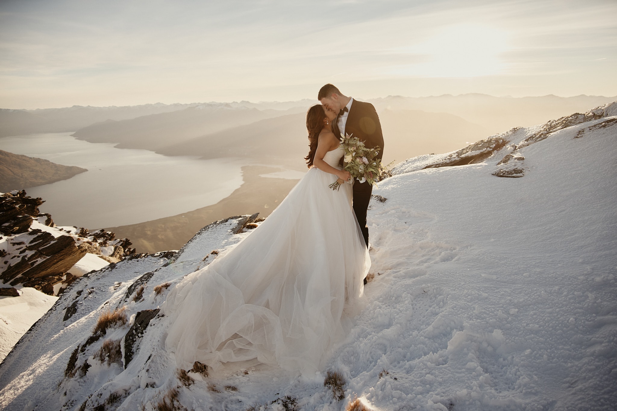 Queenstown Tekapo New Zealand Heli Wedding Elopement Pre-Wedding Shoot Photographer クイーンズタウン テカポ ニュージーランド エロープメント 前撮り フォトウェディング 結婚式 | A couple in wedding attire embracing on a snowy mountain with a lake in the background, celebrating their Queenstown Heli-Wedding & Elopement Package.