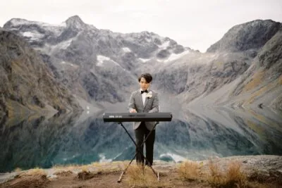 A man in a suit standing in front of a lake with mountains in the background at Coromandel Peak, Wanaka NZ.