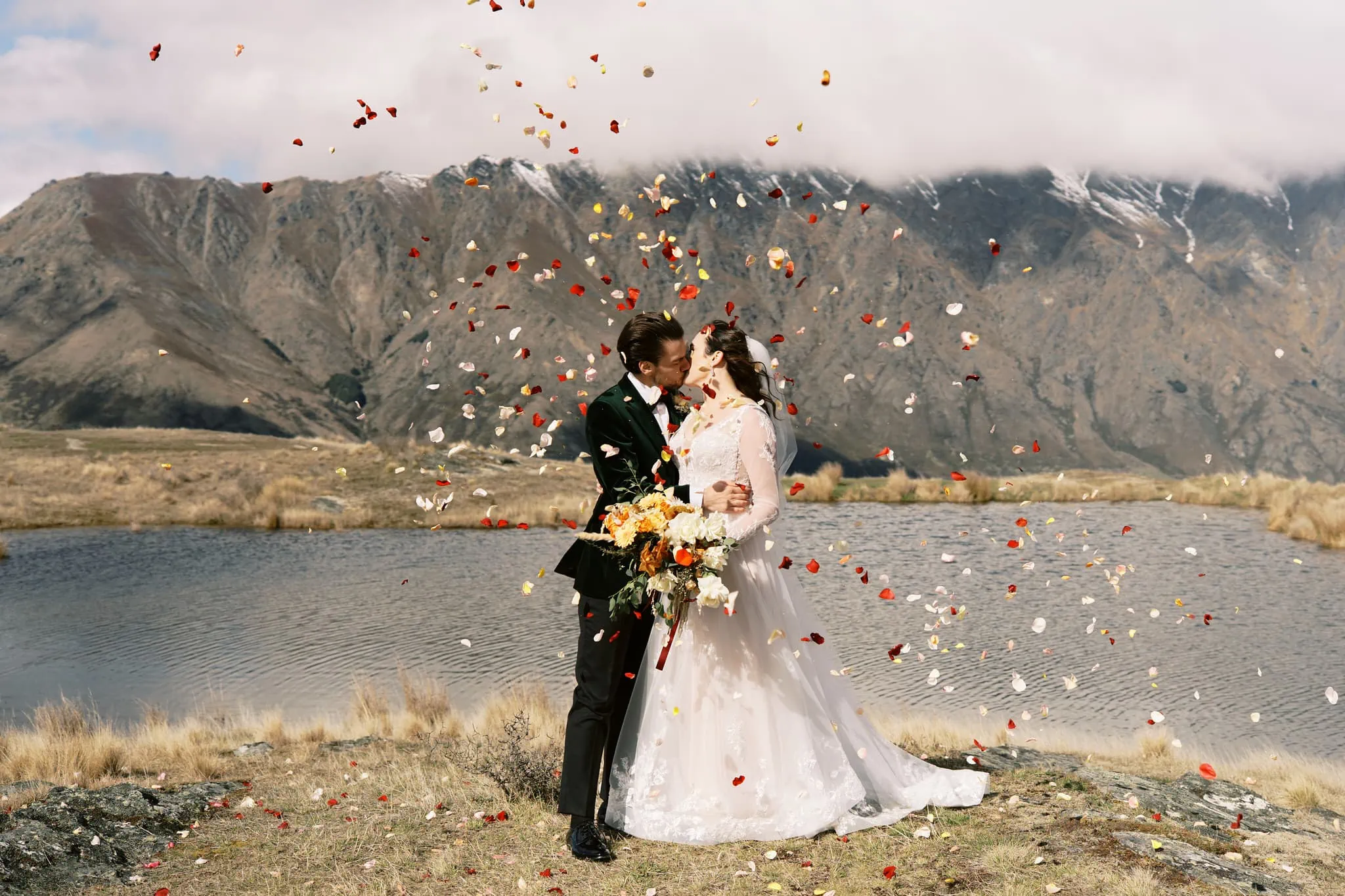Queenstown New Zealand Elopement Wedding Photographer - Josh Yates - Portfolio: A bride and groom kiss in front of a lake with confetti falling around them.