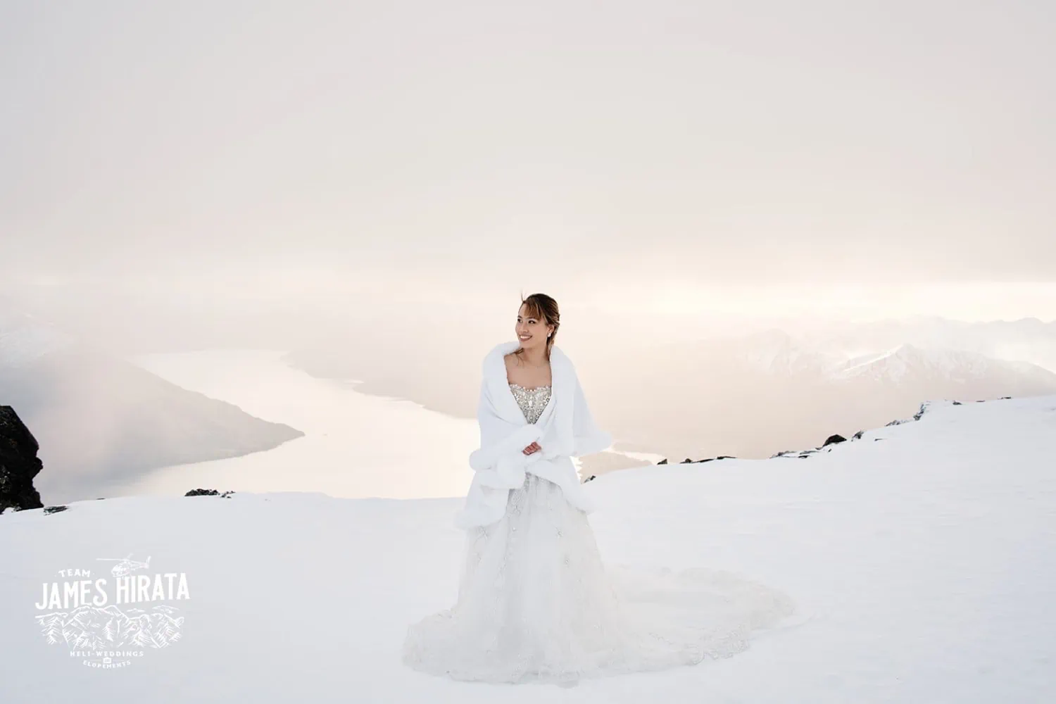 Joan & Brandon's Queenstown Heli Pre Wedding Shoot: A bride in a white dress standing on top of a snowy mountain.
