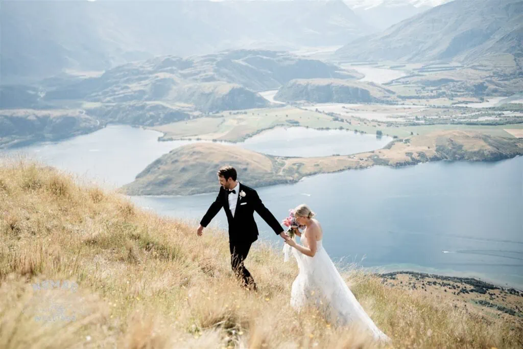 A bride and groom eloping in Queenstown, New Zealand, walking down a hill overlooking Lake Wanaka.