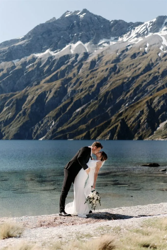An elopement wedding photographer captures a bride and groom kissing on the shore of Lake Wanaka in Queenstown, New Zealand.