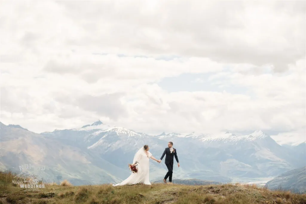 Rachel & Hamish's Coromandel Peak Heli Wedding captures a breathtaking moment of the bride and groom standing on a hill with majestic mountains in the background.