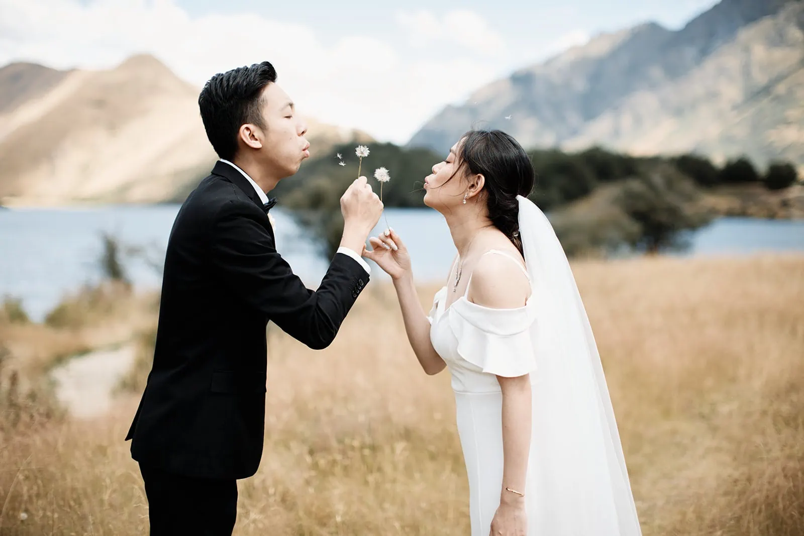 A couple blowing a dandelion in a field during their Queenstown Heli Pre Wedding at Cecil Peak.