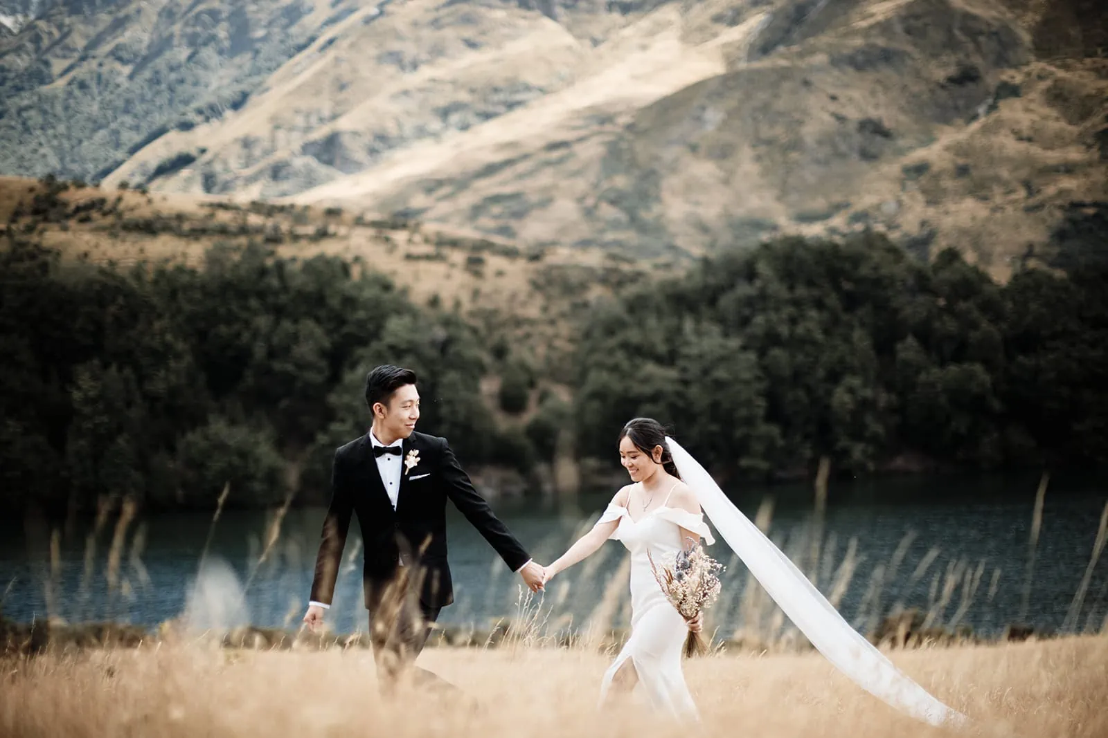 A bride and groom walking through a field with mountains in the background during their Queenstown Heli Pre Wedding at Cecil Peak.