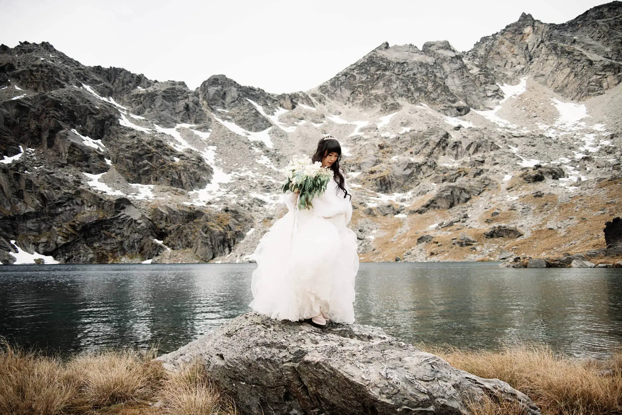 Carlos and Wanzhu's intimate heli elopement wedding at Cecil Peak with a bride standing on a rock next to a lake in the mountains.