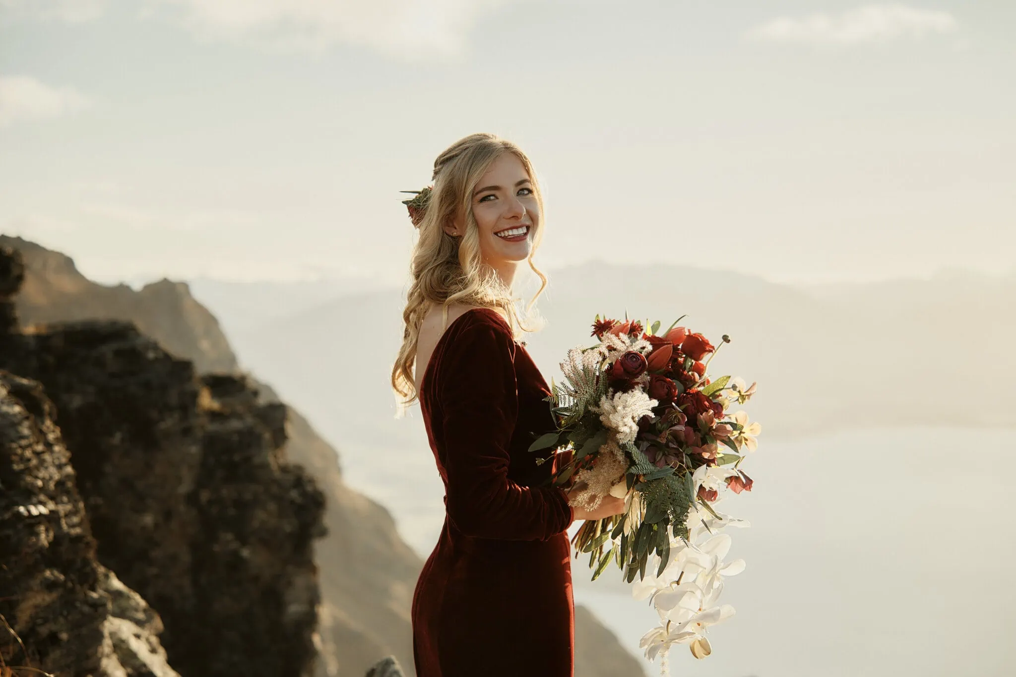 Claire and Rob's heli elopement wedding at Cecil Peak featured a stunning bride in a burgundy dress holding a bouquet on top of the mountain.