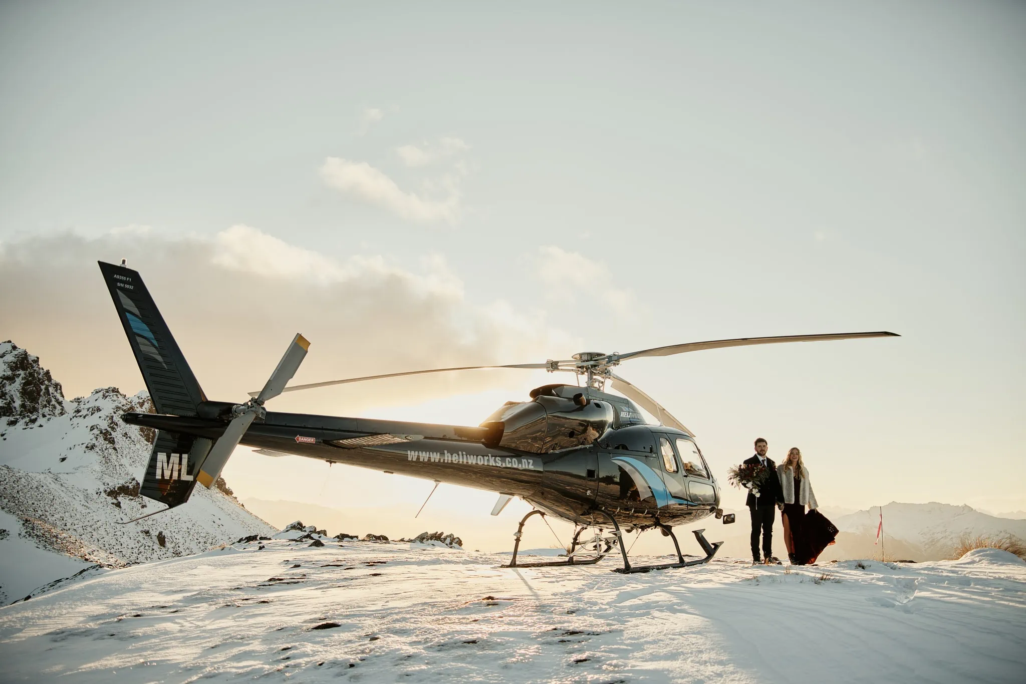 Claire and Rob posing with a helicopter in the snow during their Heli Elopement Wedding at Cecil Peak.