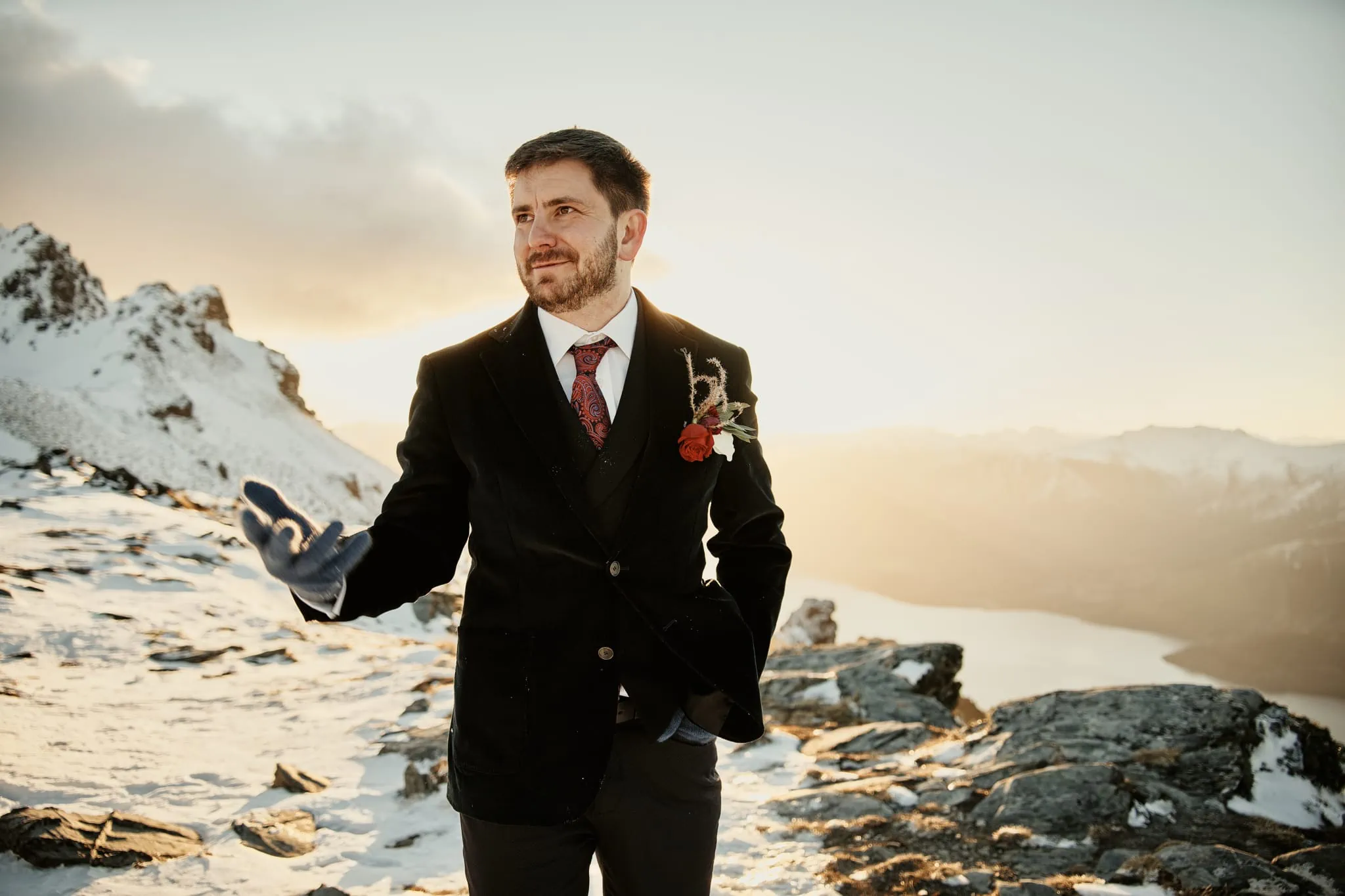 A man in a suit standing on top of a snowy mountain during Claire and Rob's heli elopement wedding at Cecil Peak.