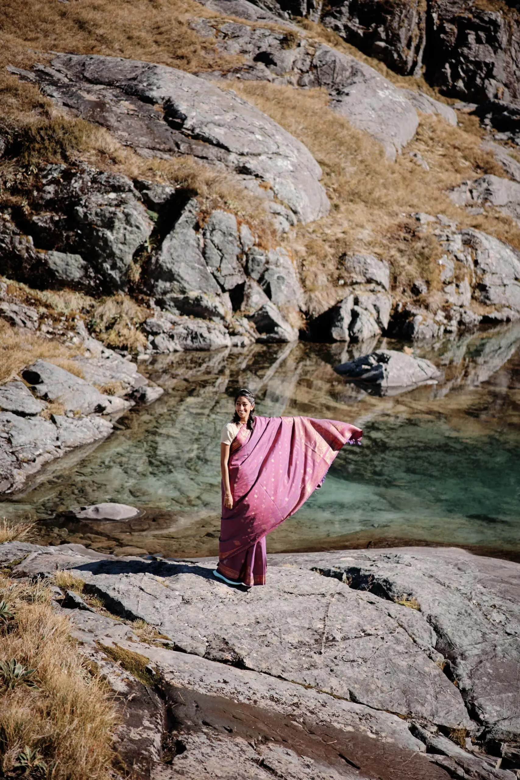 A woman in a pink shawl standing on rocks near a body of water.